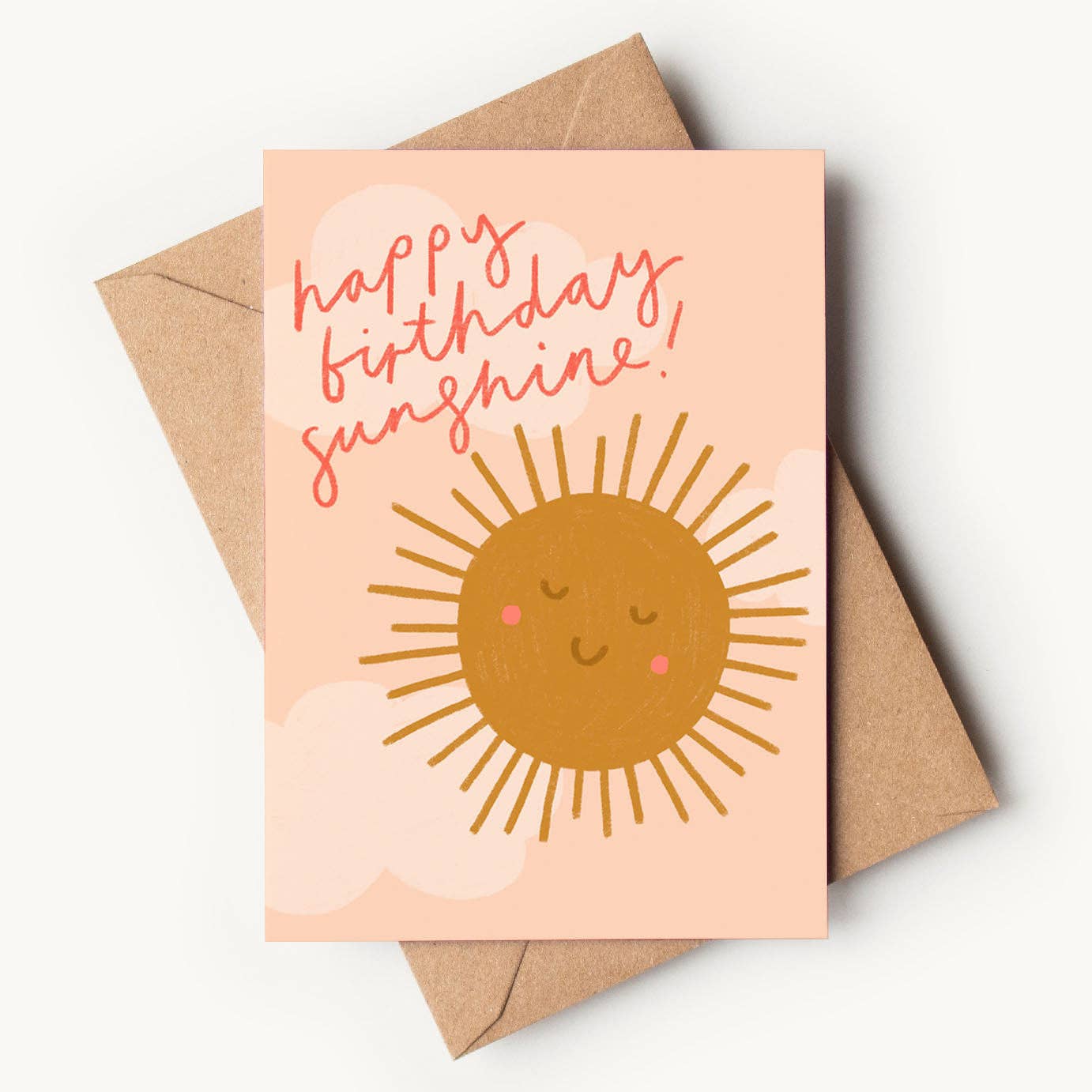 A peach-colored textured greeting card featuring a friendly, hand-drawn golden-brown sun illustration with a smiling face and pink rosy cheeks. Soft, pale pink clouds form the background, and red cursive handwriting reads "happy birthday sunshine!", resting on a brown kraft envelope.