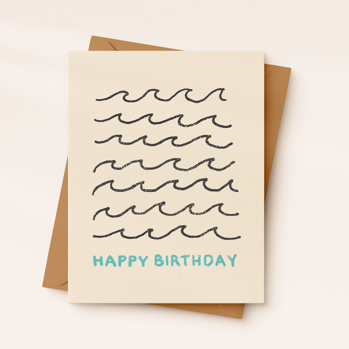 A cream-colored textured greeting card featuring seven rows of minimalist, hand-drawn black ocean wave illustrations with the text "HAPPY BIRTHDAY" printed in light blue below, resting on a brown kraft envelope.