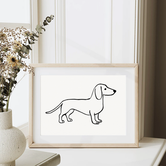 Minimalist black and white line drawing of a sausage dog (dachshund) displayed in a natural wood frame on a light surface beside a neutral ceramic vase with dried flowers. Modern Scandi-style dog wall art.