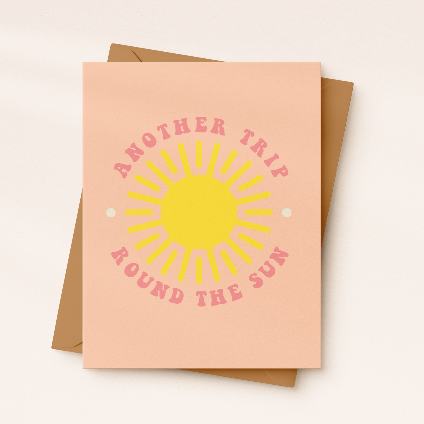 A peach-colored textured greeting card featuring a large, bright yellow graphic sun in the center. Pink retro block text curves around the sun reading "ANOTHER TRIP" at the top and "ROUND THE SUN" at the bottom, resting on a brown kraft envelope.