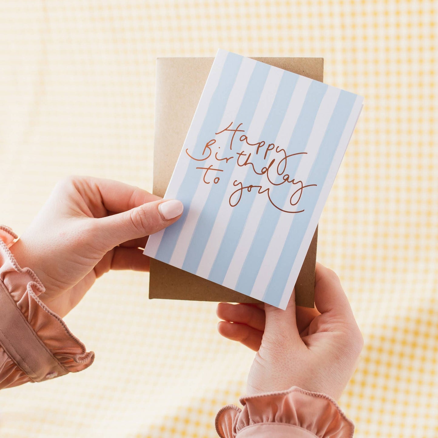 pair of hands holding a vertical greeting card featuring light blue and white vertical stripes with elegant copper foil cursive hand lettering that reads "Happy Birthday to you", backed by a kraft envelope.
