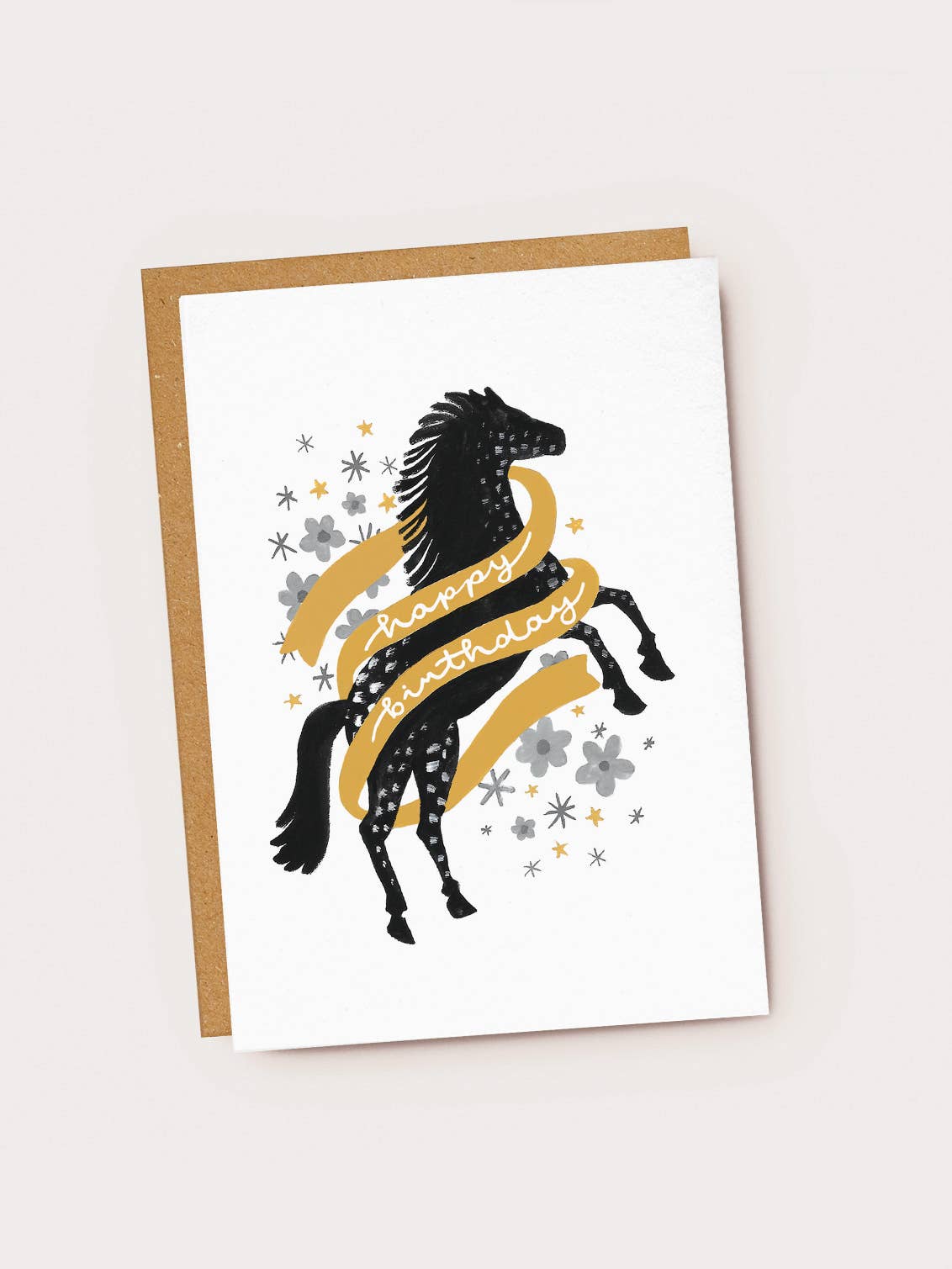 A white textured greeting card featuring a black folk-art style rearing horse with white speckled patterns on its coat. A golden-yellow banner wraps around the horse, featuring white cursive script that reads "happy birthday". The background is decorated with grey flowers, grey stars, and tiny gold stars, resting on a brown kraft envelope.