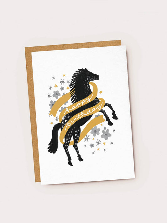 A white textured greeting card featuring a black folk-art style rearing horse with white speckled patterns on its coat. A golden-yellow banner wraps around the horse, featuring white cursive script that reads "happy birthday". The background is decorated with grey flowers, grey stars, and tiny gold stars, resting on a brown kraft envelope.