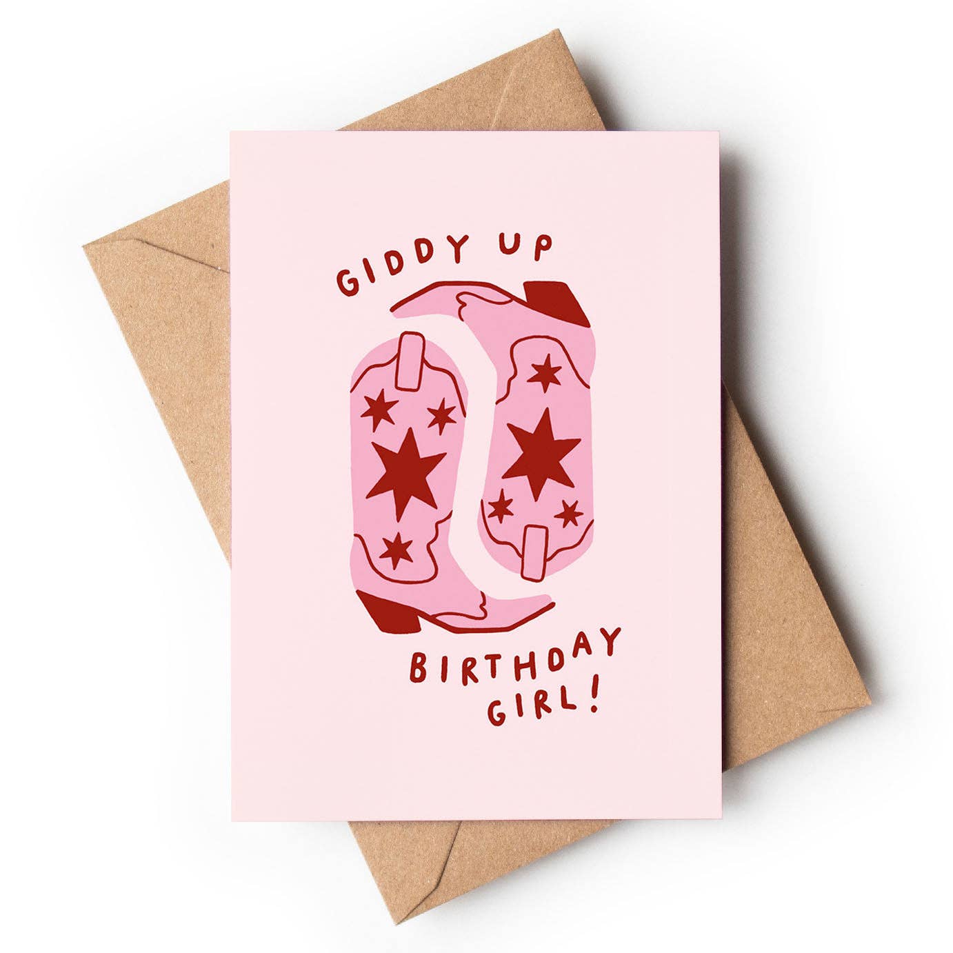 A light pastel pink textured greeting card featuring an illustration of two pink cowboy boots decorated with dark red stars. Playful red block text curves above and below the boots reading "GIDDY UP" and "BIRTHDAY GIRL!", resting on a brown kraft envelope.