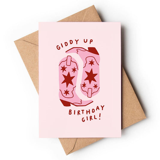 A light pastel pink textured greeting card featuring an illustration of two pink cowboy boots decorated with dark red stars. Playful red block text curves above and below the boots reading "GIDDY UP" and "BIRTHDAY GIRL!", resting on a brown kraft envelope.
