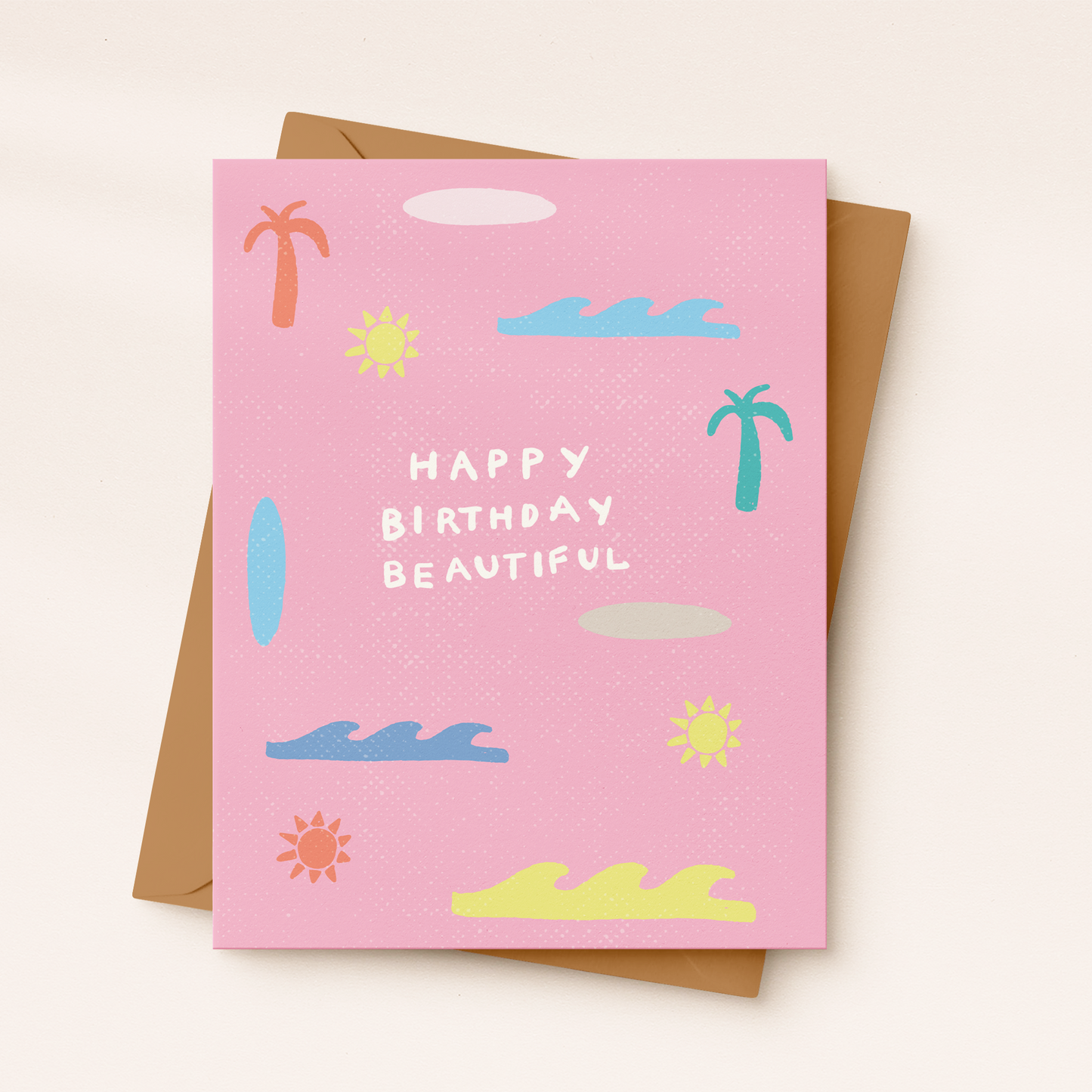 A textured pink greeting card featuring white hand-lettered text that reads "HAPPY BIRTHDAY BEAUTIFUL" surrounded by colorful beach illustrations including waves, palm trees, suns, and a surfboard, resting on a kraft paper envelope.