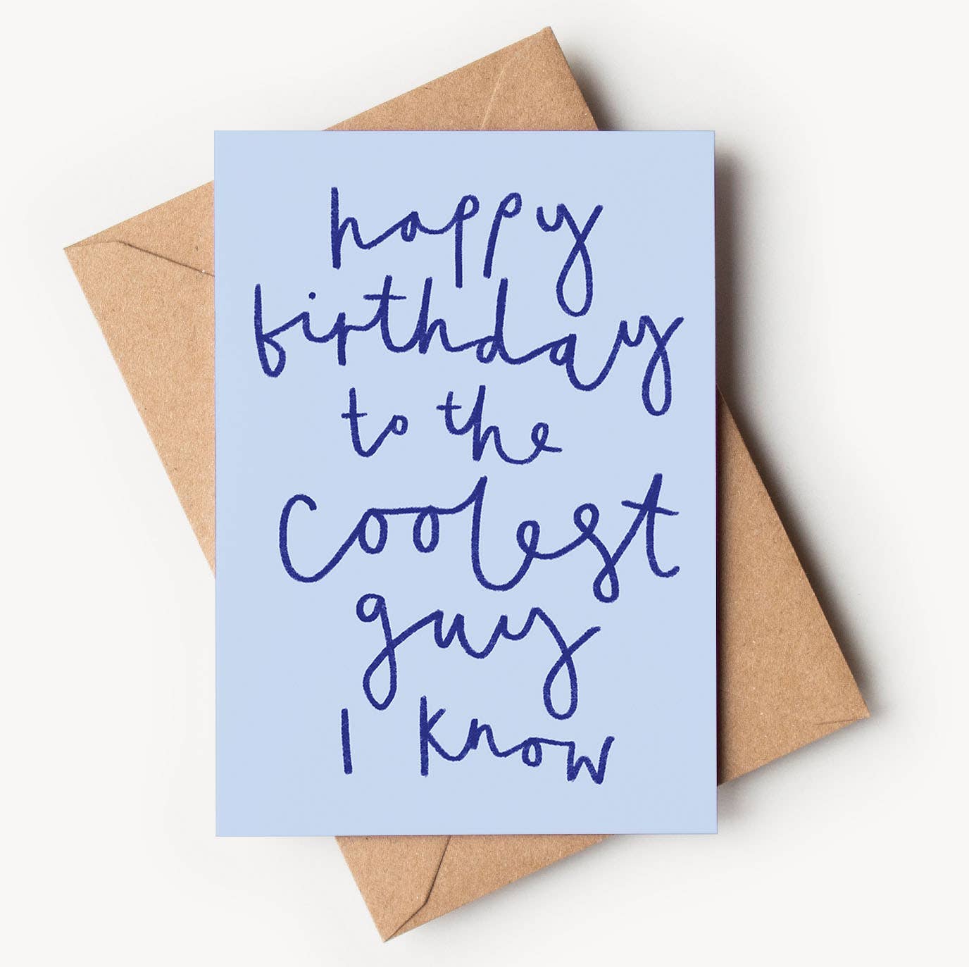 A light blue textured greeting card featuring casual, dark blue hand-written script that reads "happy birthday to the coolest guy I know", resting on a brown kraft envelope.