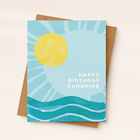 A light blue greeting card featuring a large, textured yellow watercolour sun shining over teal ocean waves, with white block text reading "HAPPY BIRTHDAY SUNSHINE", resting on a kraft paper envelope.