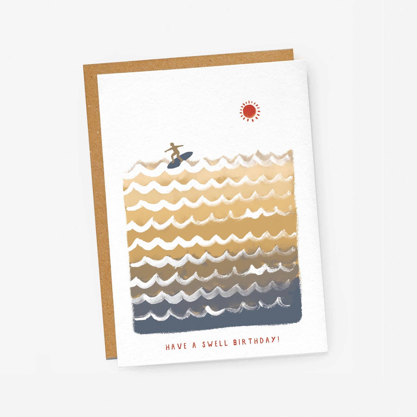 A white textured greeting card featuring a retro-style block illustration of ocean waves shifting in color from sand-gold down to deep slate blue. A tiny silhouette of a surfer rides a wave near the top under a small red sun. Red sans-serif text at the bottom reads "HAVE A SWELL BIRTHDAY!", resting on a brown kraft envelope.