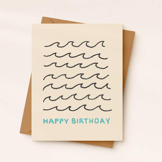 A cream-colored textured greeting card featuring seven rows of minimalist, hand-drawn black ocean wave illustrations with the text "HAPPY BIRTHDAY" printed in light blue below, resting on a brown kraft envelope.