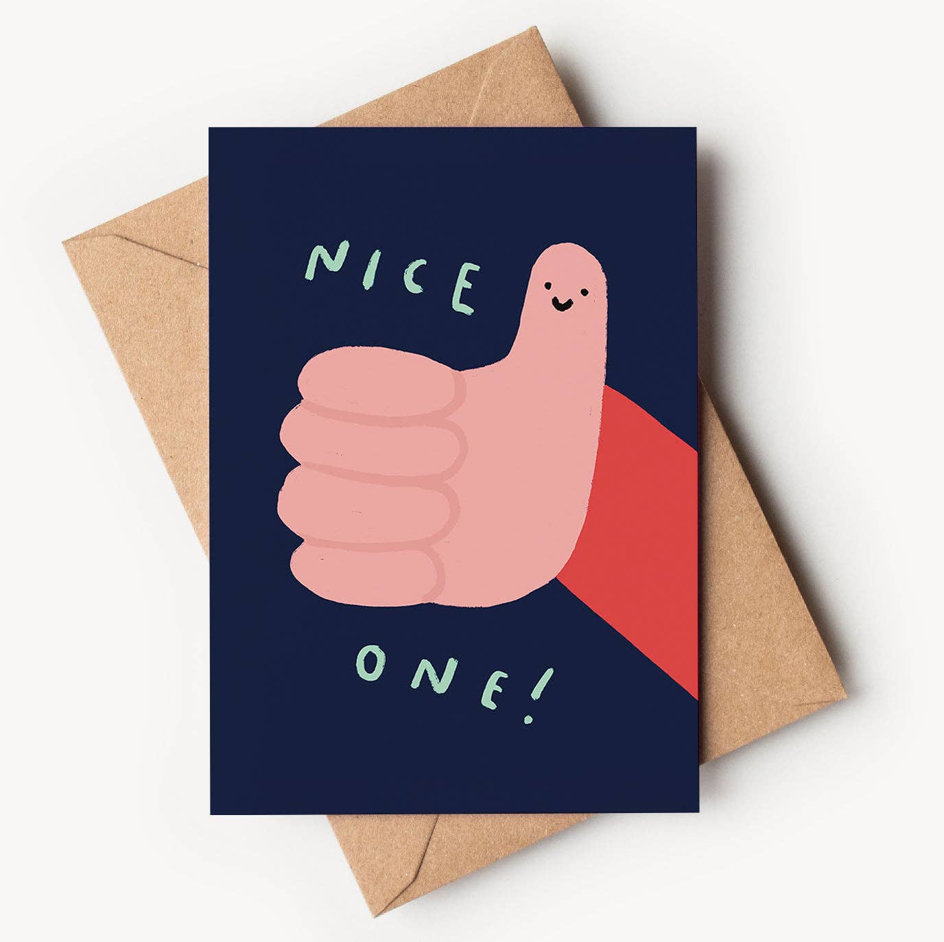 A dark navy blue textured greeting card featuring a playful illustration of a pink hand giving a big thumbs up with a tiny smiling face drawn on the thumb. Light green hand-lettered text reads "NICE" at the top and "ONE!" at the bottom, resting on a brown kraft envelope.