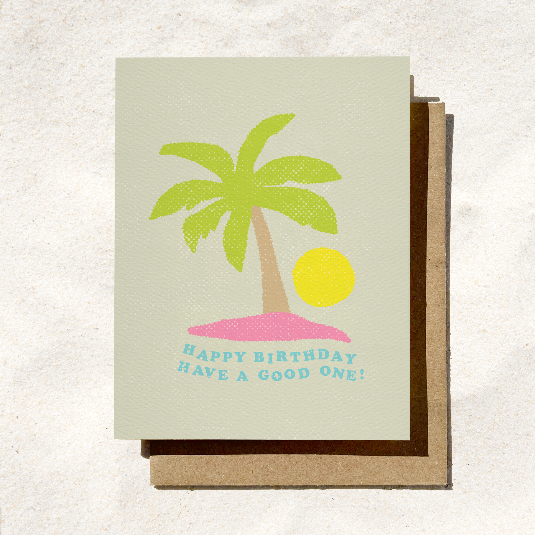 A light sage green textured greeting card featuring a stylized illustration of a green palm tree on a small pink island next to a bright yellow sun. Curved blue text underneath reads "HAPPY BIRTHDAY HAVE A GOOD ONE!", resting on a brown kraft envelope.