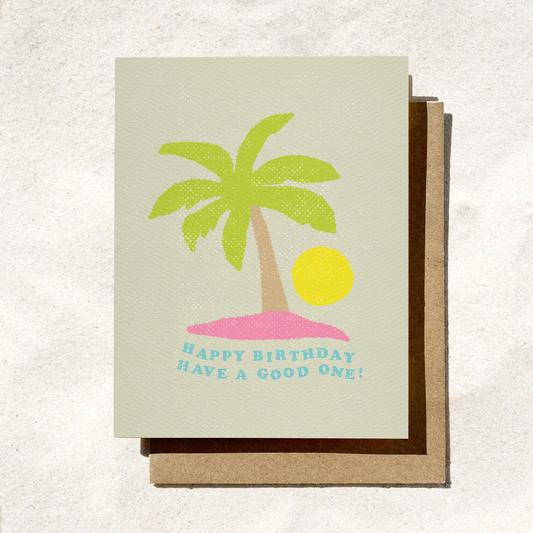 A light sage green textured greeting card featuring a stylized illustration of a green palm tree on a small pink island next to a bright yellow sun. Curved blue text underneath reads "HAPPY BIRTHDAY HAVE A GOOD ONE!", resting on a brown kraft envelope.
