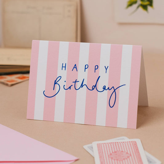 Landscape greeting card featuring soft pink and white vertical candy stripes with bold blue foil cursive hand lettering reading "Happy Birthday".
