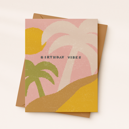 A textured greeting card featuring an abstract retro coastal landscape with a dusty pink sky, a golden yellow sun, and sand dunes. A large cream palm tree and a smaller sage green palm tree frame the center, where dark text reads "BIRTHDAY VIBES", resting on a brown kraft envelope.