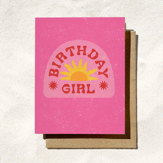 A vibrant pink textured greeting card featuring a lighter pink arch illustration in the center. Inside the arch is a yellow and orange retro sunburst graphic flanked by two red starbursts, with bold red text that reads "BIRTHDAY GIRL", resting on a brown kraft envelope.