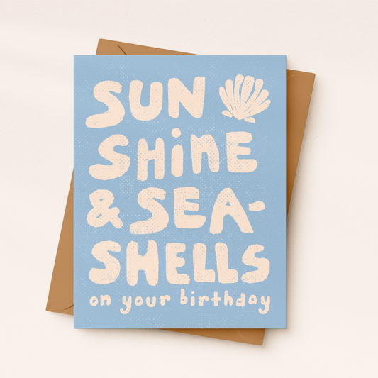 A light blue textured greeting card with cream-colored block lettering reading "SUN SHINE & SEA-SHELLS on your birthday" featuring a small seashell illustration, resting on a kraft paper envelope.