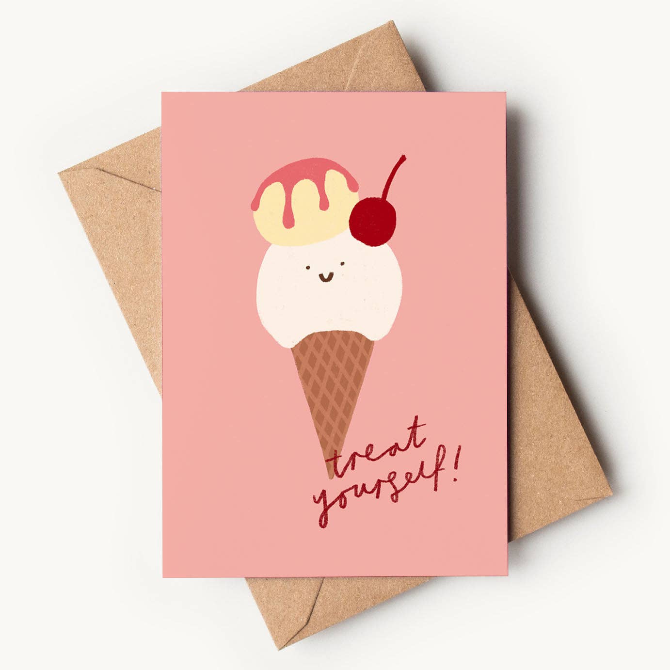 A warm pink textured greeting card featuring a cute hand-drawn waffle cone with two scoops of ice cream. The bottom scoop is white with a tiny smiling face, the top scoop is pale yellow with pink syrup dripping down, and a bright red cherry sits on the side. Red cursive text at the bottom reads "treat yourself!", resting on a brown kraft envelope.
