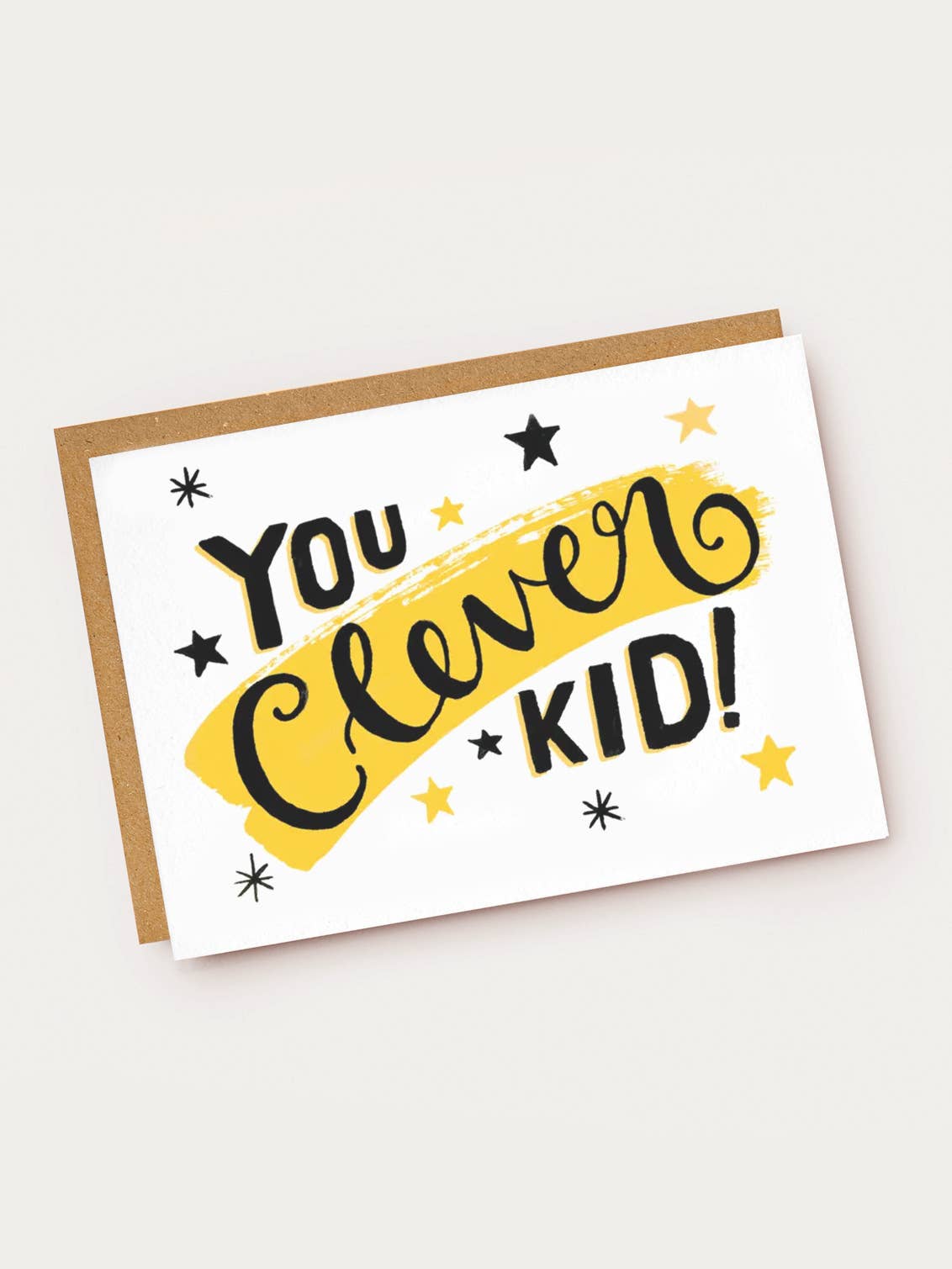 A white landscape-oriented textured greeting card featuring bold hand-lettered text that reads "YOU Clever KID!". The word "Clever" is written in elegant cursive over a bright yellow brushstroke background. The card is decorated with black and yellow stars and sparkles, resting on a brown kraft envelope.