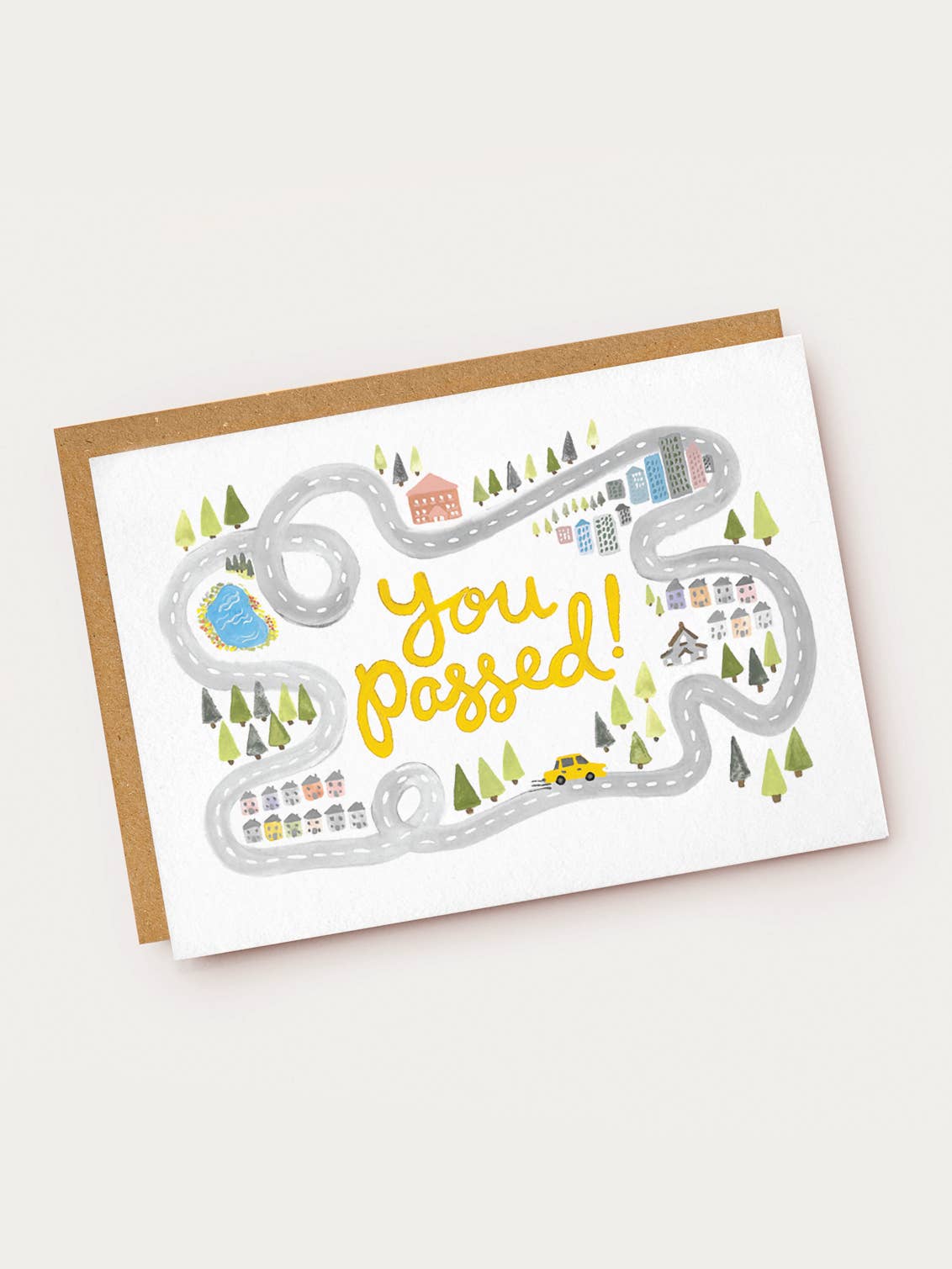 A white landscape-oriented textured greeting card featuring a watercolor illustration of a winding gray road with loops, tiny houses, pine trees, a blue pond, and a small yellow car. Bright yellow hand-lettered text in the center reads "you passed!", resting on a brown kraft envelope.