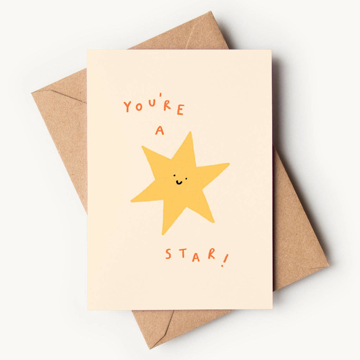 A cream-colored textured greeting card featuring a simple, hand-drawn yellow star with a cute smiling face in the center. Hand-lettered orange text reads "YOU'RE A" at the top and "STAR!" at the bottom, resting on a brown kraft envelope.