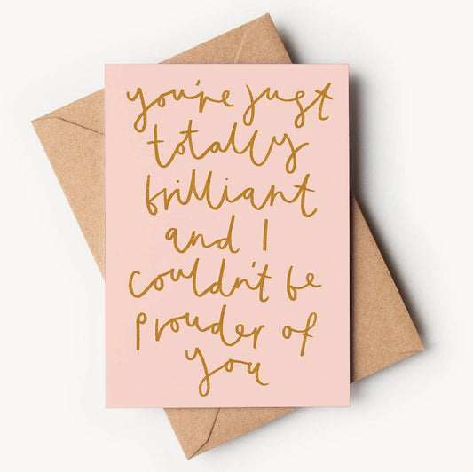 A soft pastel pink textured greeting card featuring elegant, golden-brown handwritten script that fills the front, reading "you're just totally brilliant and I couldn't be prouder of you", resting on a brown kraft envelope.